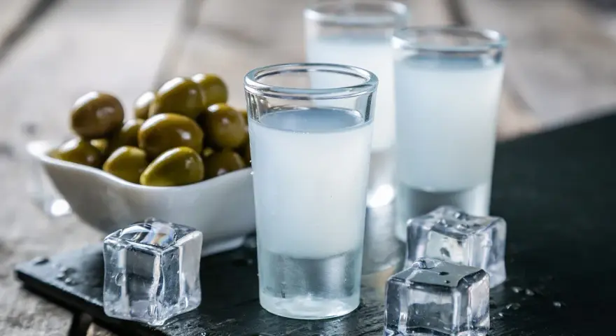 Trzy kieliszki ouzo z lodem i miseczka oliwek na drewnianym tle.