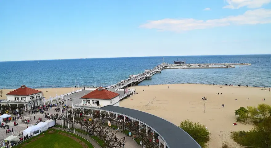Sopot beach z molo, kawiarniami i spacerującymi ludźmi. W oddali widać statki.