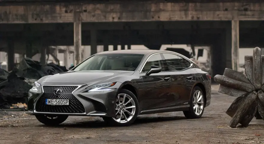 Szary Lexus LS 500h zaparkowany na tle zniszczonej budowli.
