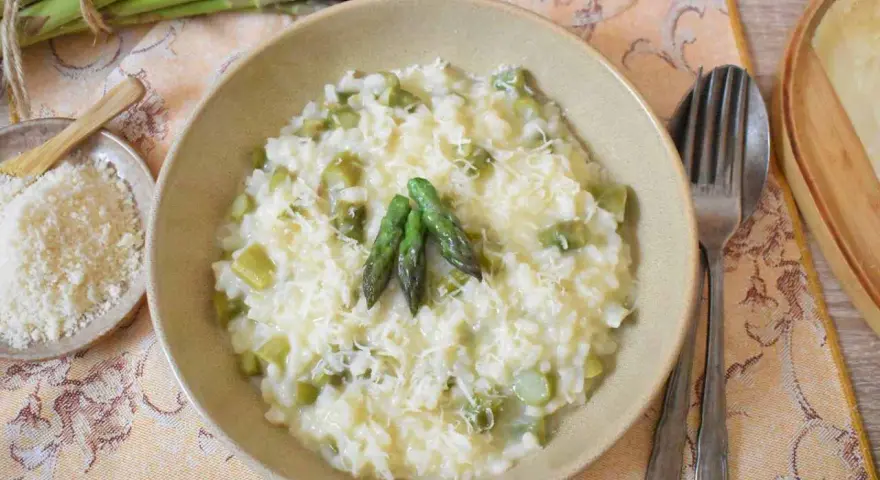Kremowe risotto ze szparagami, posypane parmezanem, z trzema zielonymi szparagami na wierzchu. Obok miseczka z tartym serem i pęczek świeżych szparagów.