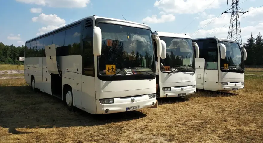 Trzy białe autokary, jeden z otwartymi drzwiami, stoją na trawie pod błękitnym niebem. Na przedniej szybie jednego z nich widnieje znak "autobus 5".
