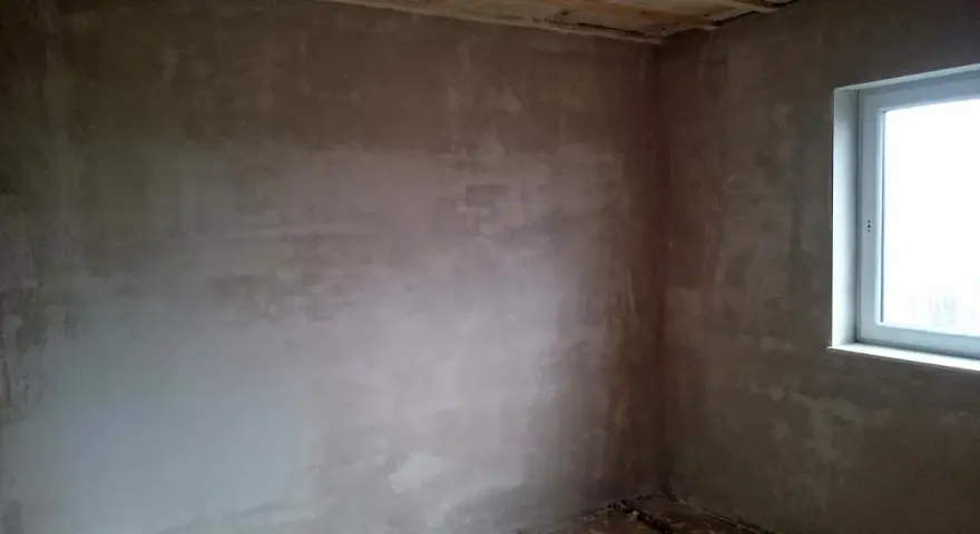 Unverputzte Innenwand mit Fensterecke und sichtbaren Deckenbalken.