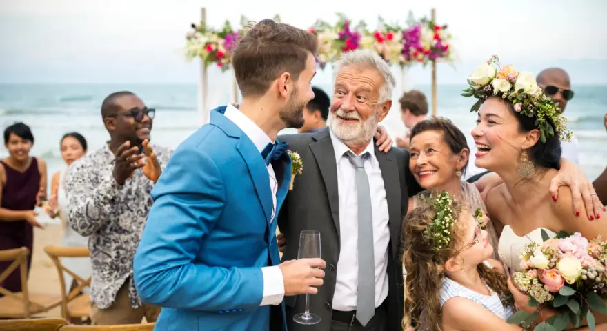 Szczęśliwy ojciec pana młodego w eleganckim garniturze śmieje się z synem na plażowym weselu.