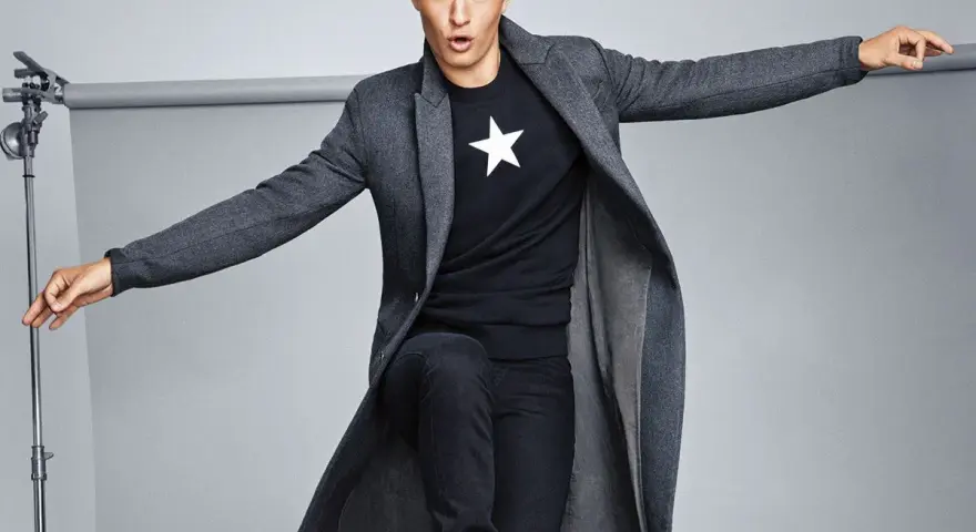 Joven con abrigo largo gris, camiseta negra con estrella blanca y vaqueros oscuros. Explora diversos tipos de abrigos para tu estilo.