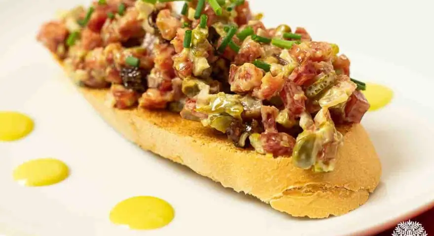 Tosta con steak tartar de salchichón y cebollino picado, decorada con puntos de salsa amarilla.