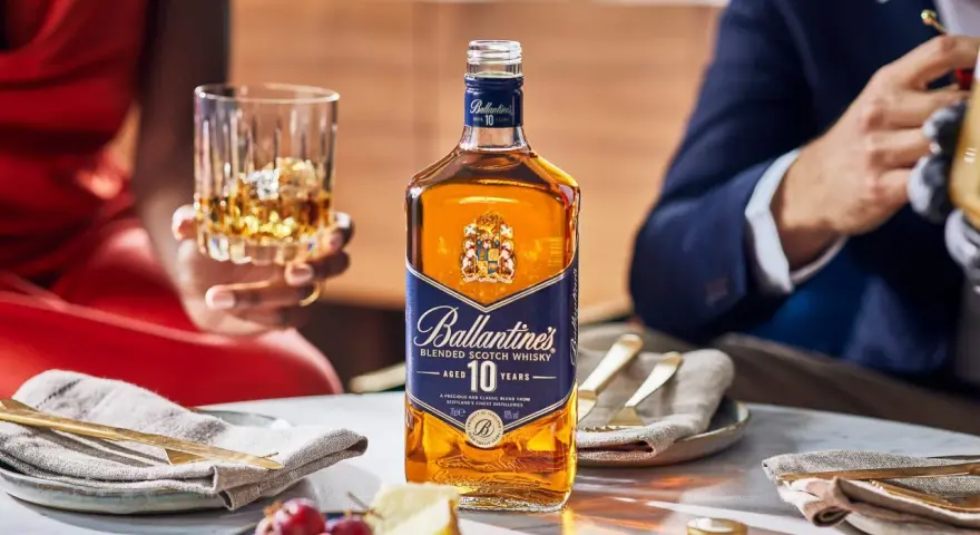 Elegancka butelka Ballantine's 10 Years Old na stole, obok kieliszka z bursztynowym płynem. To nie bourbon, lecz szkocka whisky, idealna na specjalne okazje.