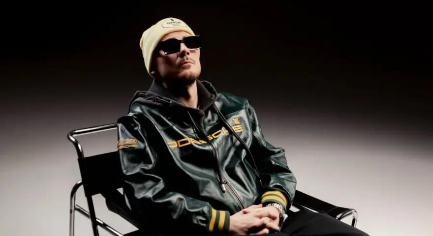 Pezet w skórzanej kurtce Porsche, w bejsbolówce i okularach, siedzi na krześle. Zapowiada nowy album.