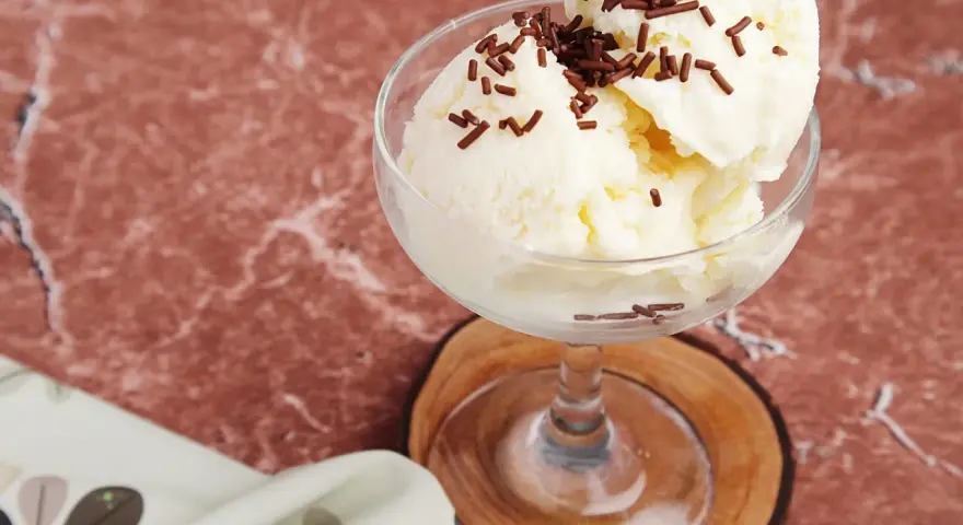 Cremiges Litschi Eis mit Schokostreuseln in einem Glas auf einem Holzuntersetzer.