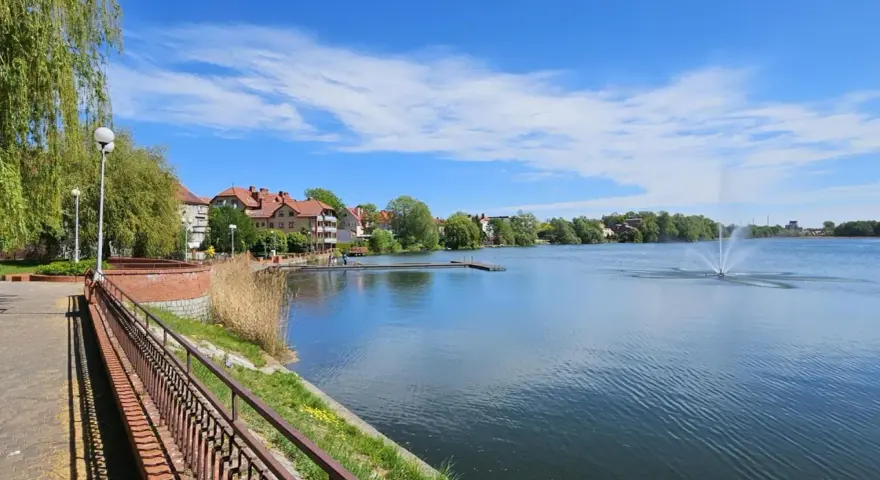Spokojne jezioro zamkowe z fontanną i drewnianym pomostem, otoczone zielenią i zabudowaniami.