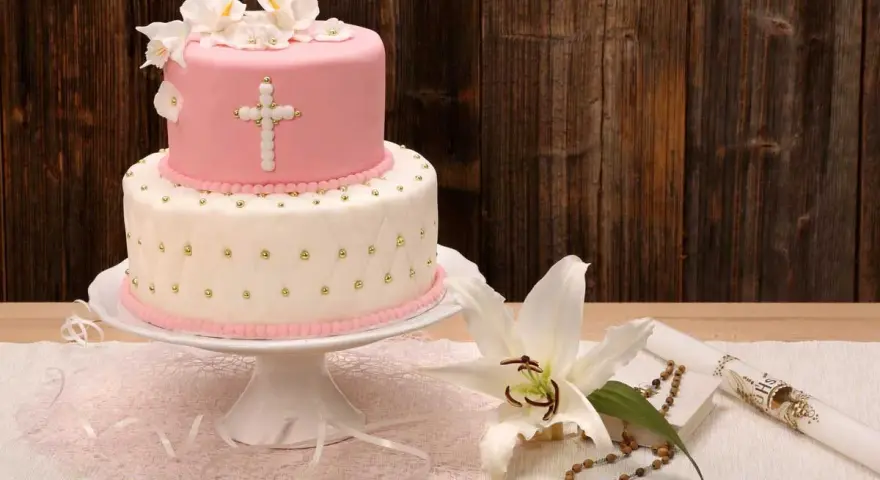 Elegancki dwupoziomowy tort na chrzest w różowo-białych barwach, ozdobiony krzyżem i kwiatami. Obok lilia, różaniec i świeca.