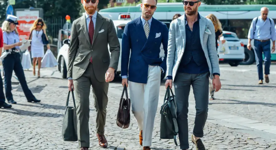 Trois hommes élégants déambulent. L'un porte une tenue avec blazer bleu ciel, un autre un costume vert olive, et le troisième un blazer gris clair.