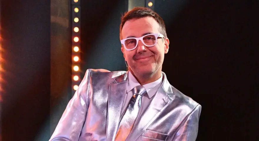 DJ Adamus w srebrnym garniturze i białych okularach, uśmiecha się. Czy to jego odpowiedź na pytanie "Jaka to melodia?