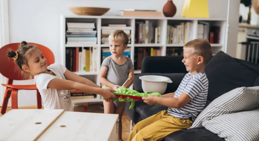 Zwei Kinder im **Streit mit Schwester** um ein Stofftier. Ein kleiner Junge schaut zu.