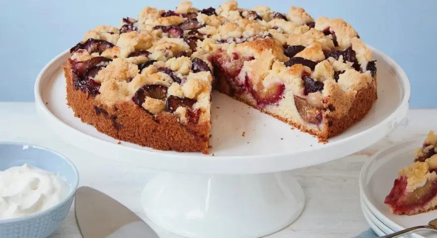 Ein einfacher Pflaumenkuchen mit Streuseln auf einem Tortenständer, daneben eine Schüssel Schlagsahne und ein Tortenheber.