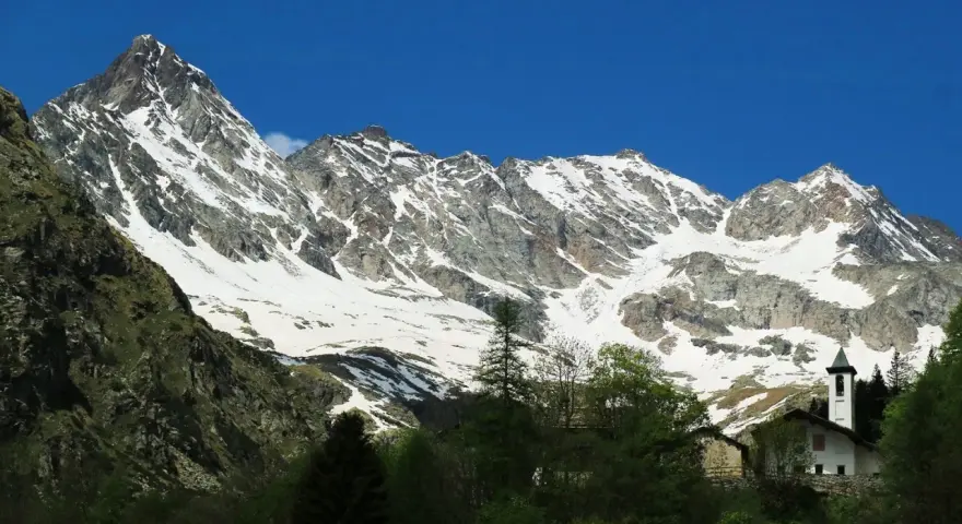 Imponente montagna sacra innevata, con un piccolo campanile bianco che emerge tra gli alberi verdi in primo piano.