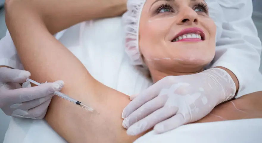 Kobieta z uśmiechem na twarzy poddaje się zabiegowi botox pod pachy. Lekarz w rękawiczkach wykonuje zastrzyk.