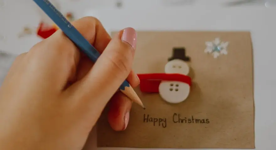 Ręka z ołówkiem pisze "Happy Christmas" na kartce z bałwankiem z guzików. Idealny przykład, jak narysować kartkę świąteczną.