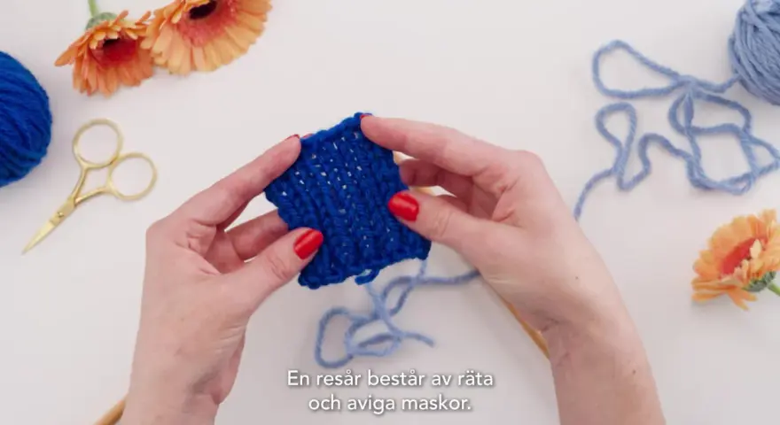 Händer som håller i ett stickat provstycke i resårstickning. Blått garn, sax och blommor syns i bakgrunden.