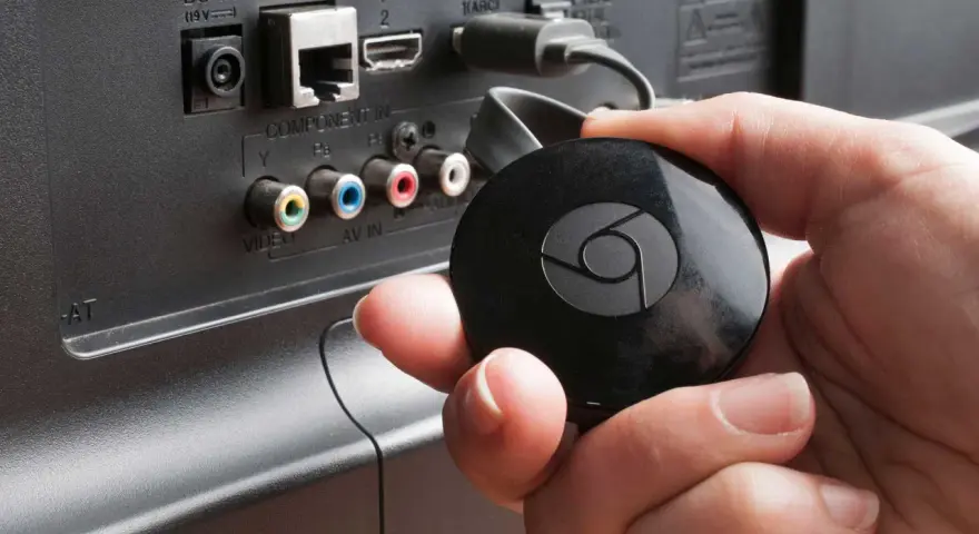 En hand håller en svart Chromecast för att ansluta till en HDMI-port på baksidan av en TV.