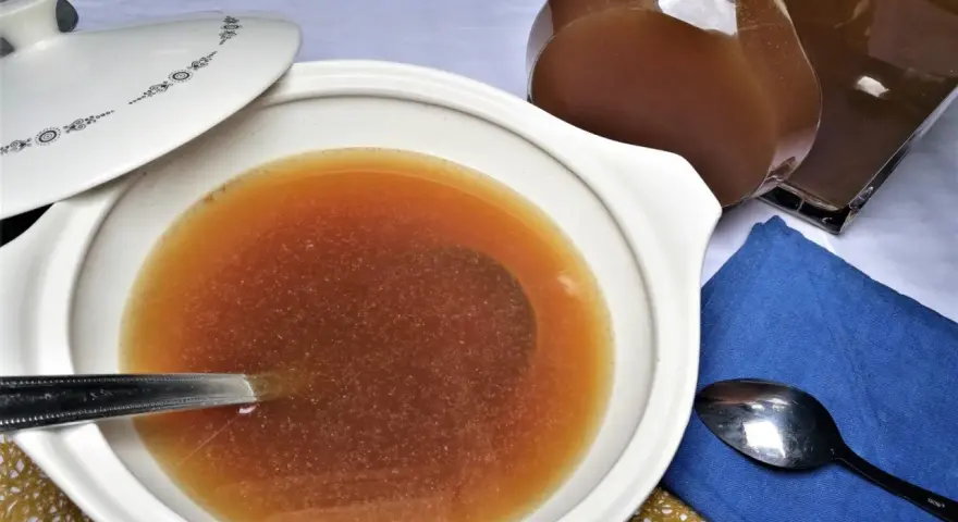 Caldo de carne casero en una cazuela blanca con tapa, junto a una jarra y cucharas.
