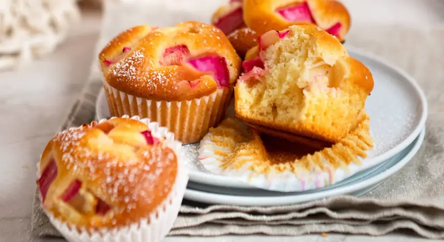 Saftige Rhabarber Muffins, mit Puderzucker bestäubt, auf einem Teller gestapelt. Einer ist angeschnitten und zeigt die rosa Rhabarberstücke.