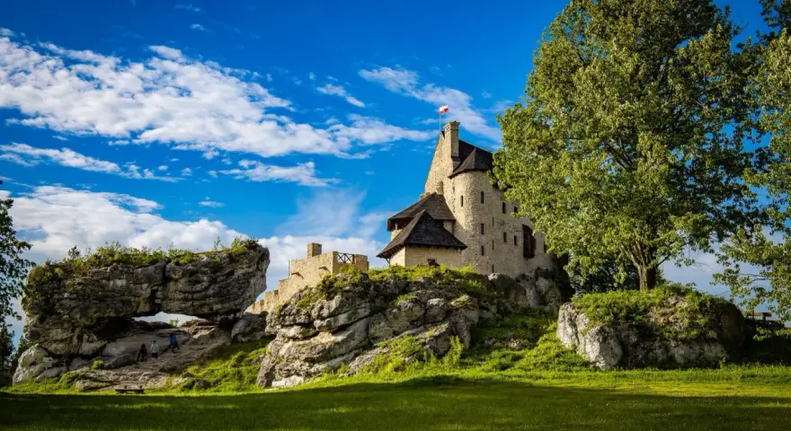 Zamek Bobolice na skale, z polską flagą, pod błękitnym niebem z chmurami. Obok skalny most i zielona trawa.