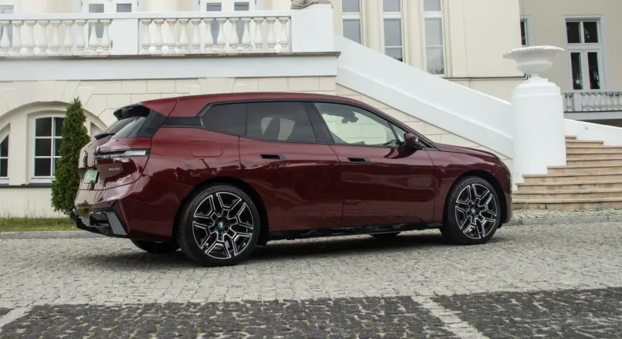 Ciemnoczerwone BMW iX stoi na brukowanym podjeździe przed eleganckim budynkiem.