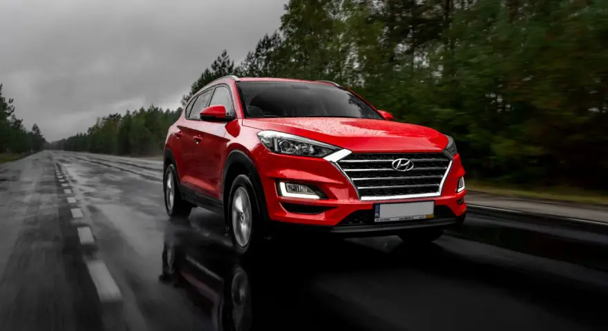Czerwony Hyundai Tucson jedzie po mokrej drodze w deszczowy dzień. Warto pamiętać o filtrze CPL, którego zastosowanie poprawia widoczność w takich warunkach.