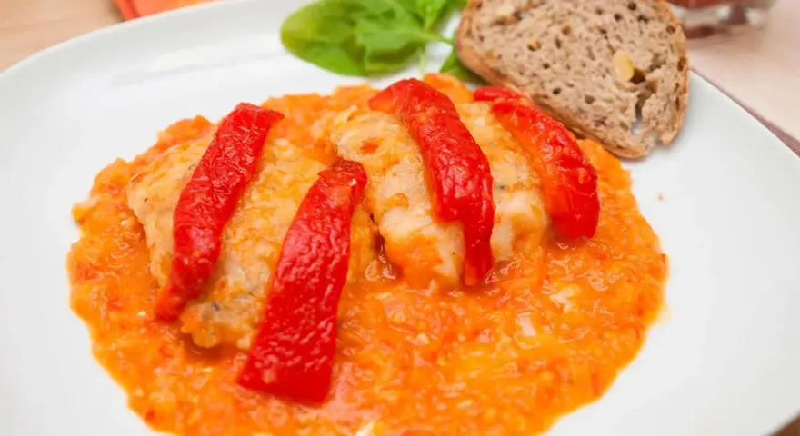 Bacalao a la riojana, un plato tradicional con trozos de pescado blanco cubiertos de pimientos rojos y una salsa de tomate y pimientos. Servido con pan y hojas verdes.