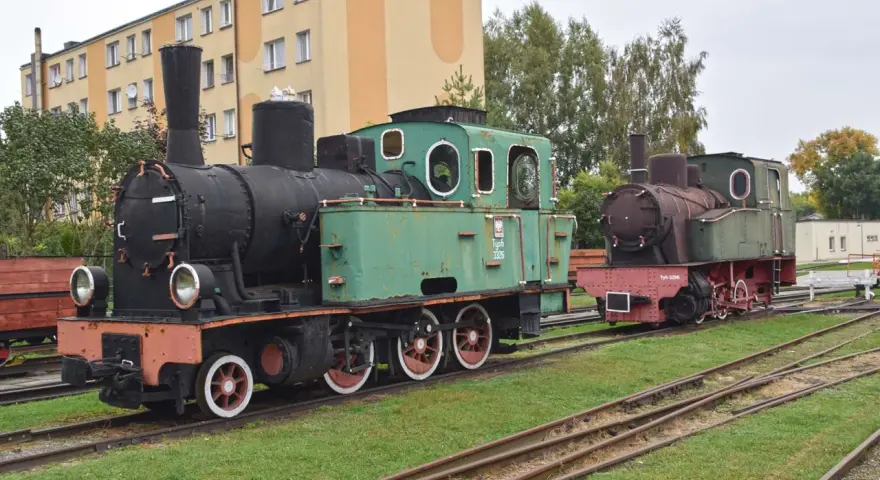 Dwa zabytkowe parowozy, zielony i rudy, stoją na torach przy budynku. To część ekspozycji muzeum kolei wąskotorowej w Sochaczewie.