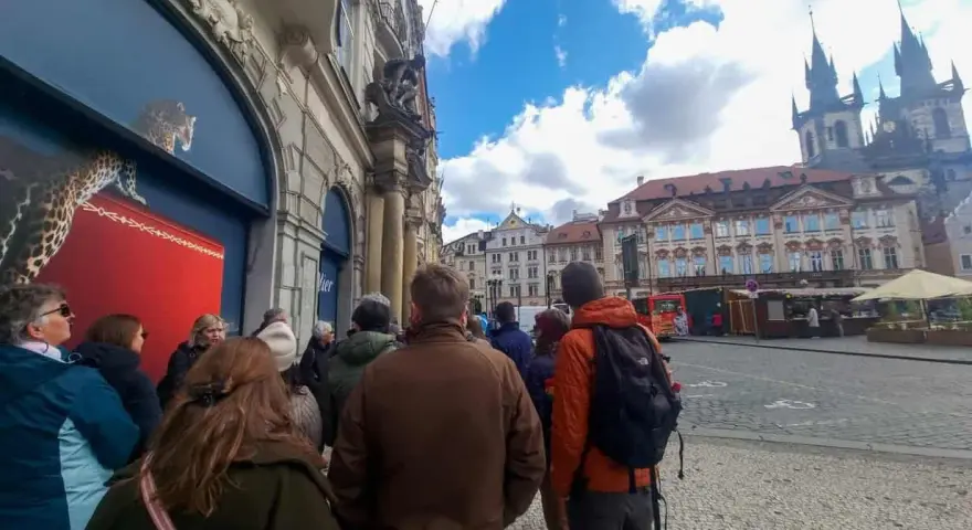 A group of tourists on a prague free walking tour gather on a cobblestone street, with historic buildings and a church in the background.