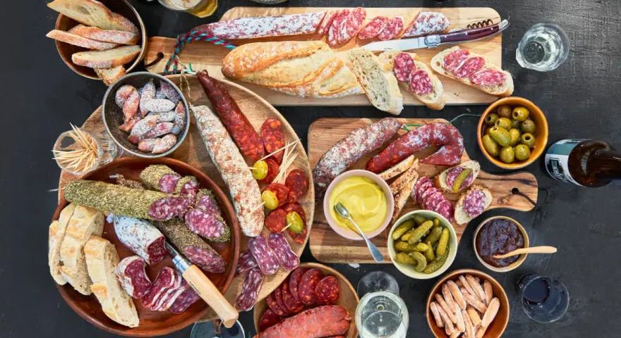 Eine reichhaltige Wurstplatte anrichten mit Baguette, Oliven, Cornichons und Senf.