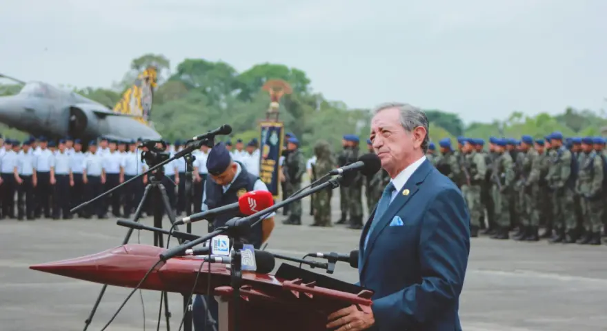 Elegancki garnitur to dobry wybór, gdy zastanawiasz się, jak się ubrać na przysięgę wojskową. Mężczyzna w granatowym garniturze przemawia na tle żołnierzy i samolotu.
