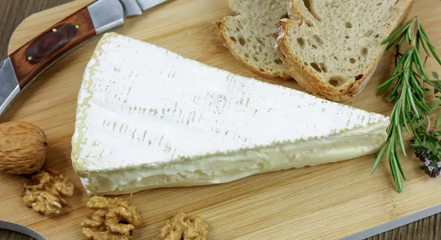Tranche de fromage à croute lavée crémeux, accompagnée de pain, noix et herbes aromatiques sur une planche en bois.