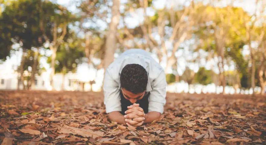 A man in a light blue shirt is in a posture of christian prostration, hands clasped on the ground covered in fallen leaves.