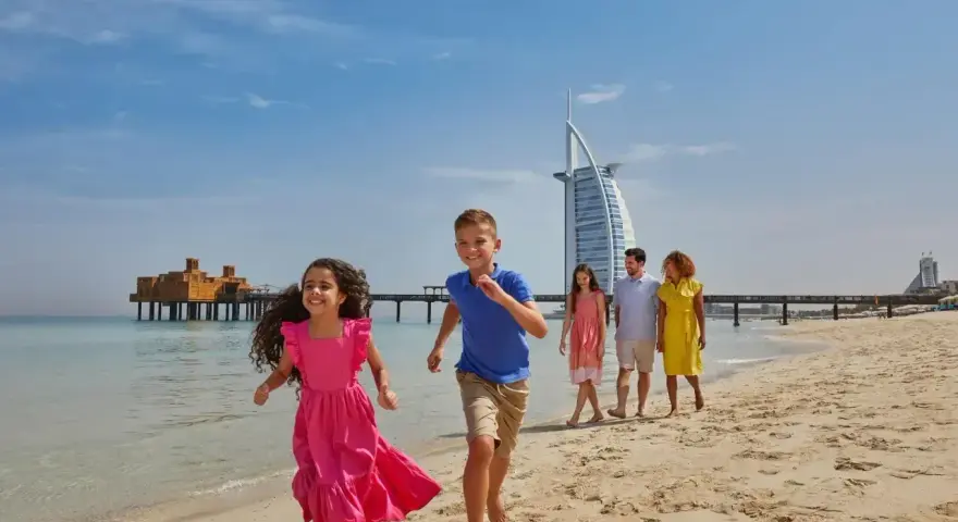 Rodzina cieszy się słonecznym dniem na plaży w Dubaju. Dzieci biegną w stronę morza, a w tle widać słynny hotel Burj Al Arab.