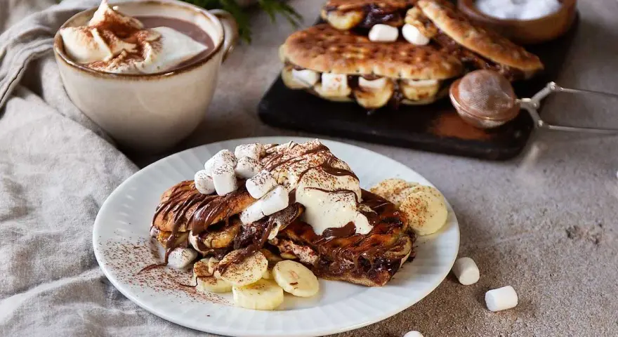 En härlig nutella macka med banan och marshmallows, toppad med grädde och kakao. Bredvid står en kopp varm choklad.