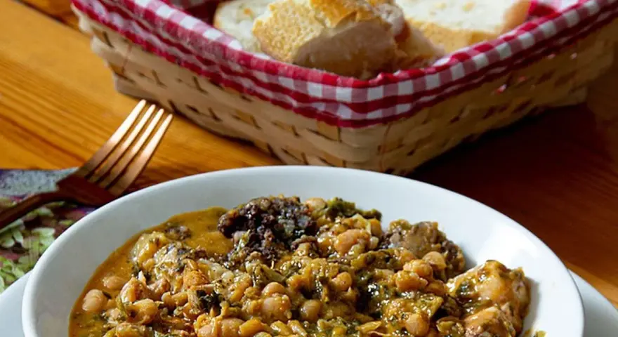 Un reconfortante cocido montañés, con garbanzos, verduras y trozos de carne, servido en un cuenco blanco. A su lado, una cesta con pan y un tenedor.