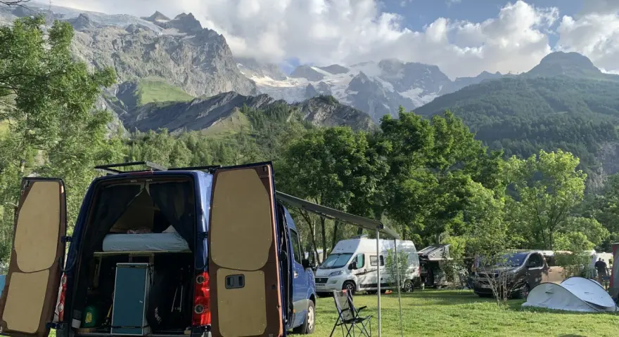 En campingplats med flera husbilar, inklusive en blå med öppna bakdörrar, i en dal med snöklädda berg i bakgrunden. En perfekt plats för äventyr med edins husbilar.