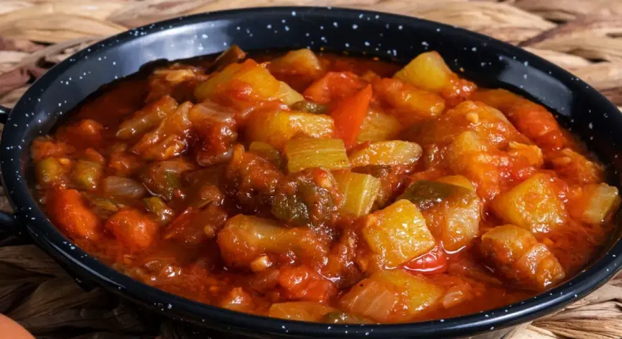 Un plato humeante de pisto manchego, con trozos de calabacín, pimiento y tomate en una salsa rojiza, servido en una cazuela negra.