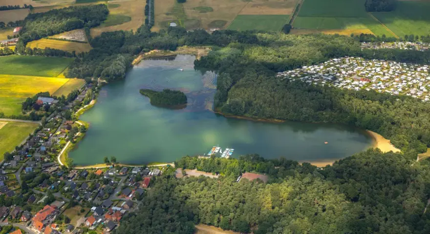 Luftaufnahme des Feldmarksees Sassenberg mit einer kleinen Insel, Booten am Steg, einem Wohngebiet und Feldern.