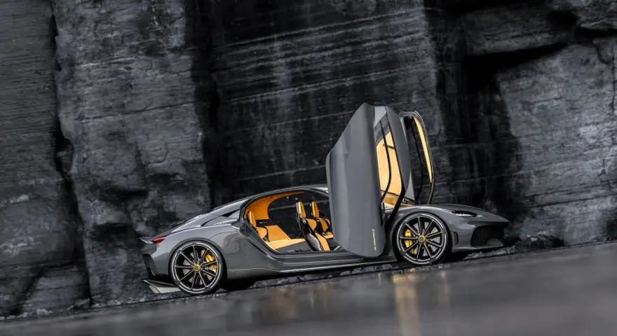 Szary Koenigsegg Gemera, czteromiejscny supersamochód z otwartymi drzwiami, prezentuje swoje żółte wnętrze na tle skalistej ściany.