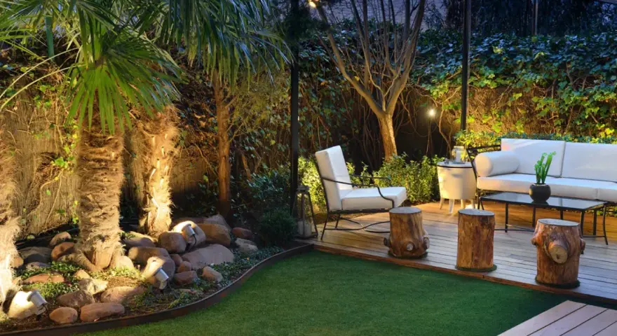 Un jardín poco mantenimiento con césped artificial, palmeras iluminadas y una zona lounge con sofá blanco y sillón.