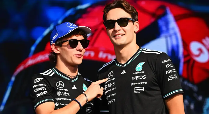 Dos pilotos de Fórmula 1, George Russell y un compañero, sonríen con gafas de sol. Russell lleva una gorra azul y ambos visten camisetas negras con logotipos de patrocinadores.