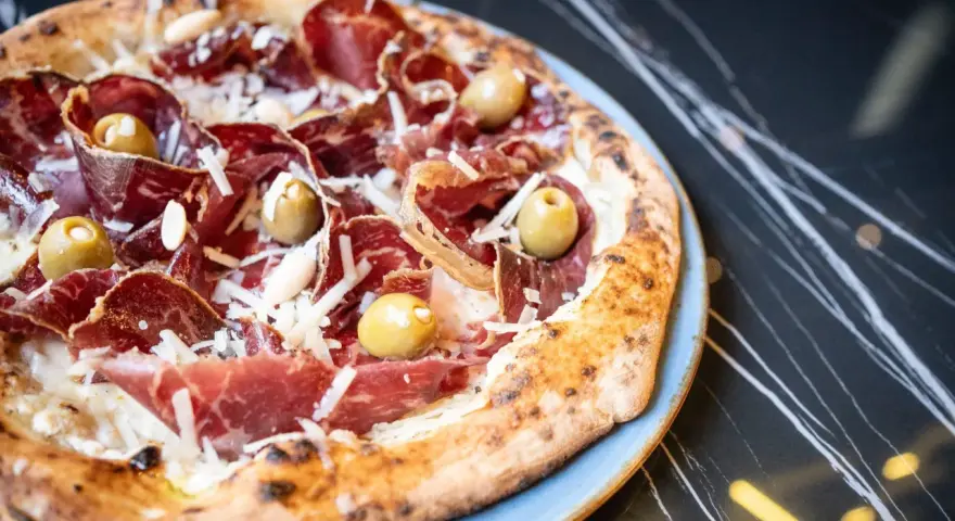 Pizza con jamón serrano, aceitunas y queso rallado, perfecta para saber donde comer en Menorca.