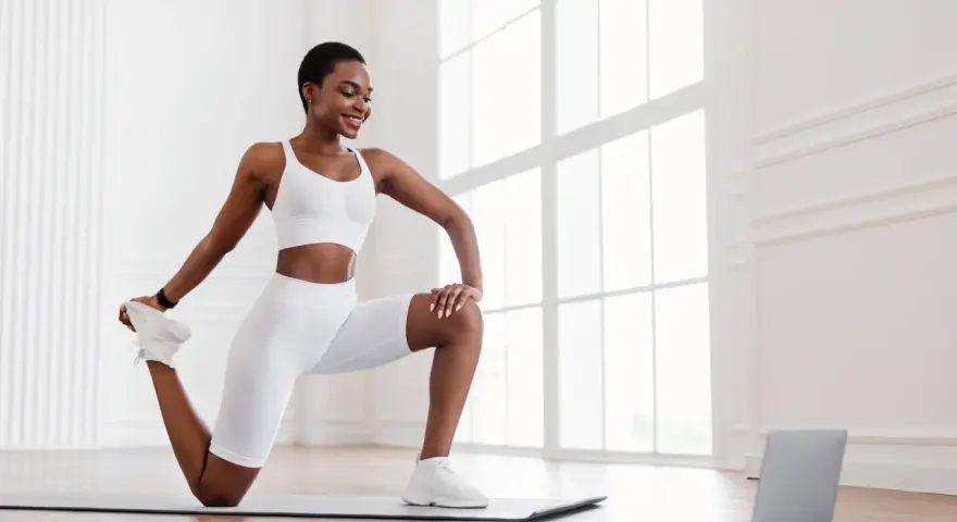 Femme en tenue de sport blanche effectuant un exercice quadriceps sur un tapis, guidée par un ordinateur portable.