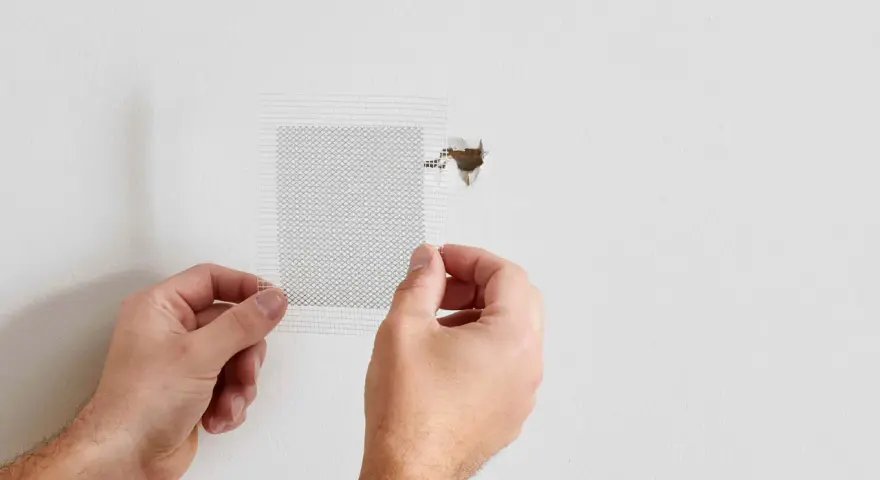 Hands apply a mesh patch to a hole in a white wall, demonstrating how to patch a hole in the wall.