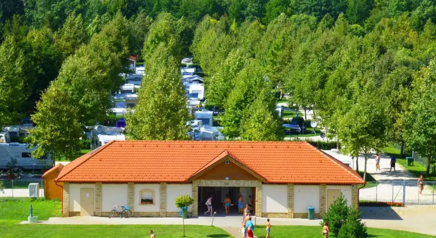 Słoneczny dzień na kempingu Liptot. Ludzie odpoczywają na trawie, a w tle widać rzędy kamperów i przyczep kempingowych wśród drzew.