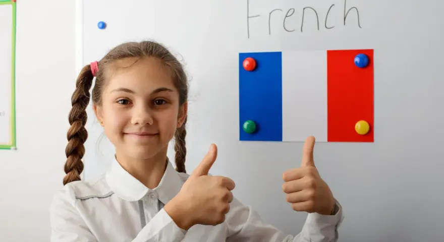 Uśmiechnięta dziewczynka z warkoczami pokazuje kciuki w górę na tle tablicy z napisem "French" i flagą Francji. To symbol pozytywnego podejścia do nauki, być może w ramach systemu edukacji we Francji.