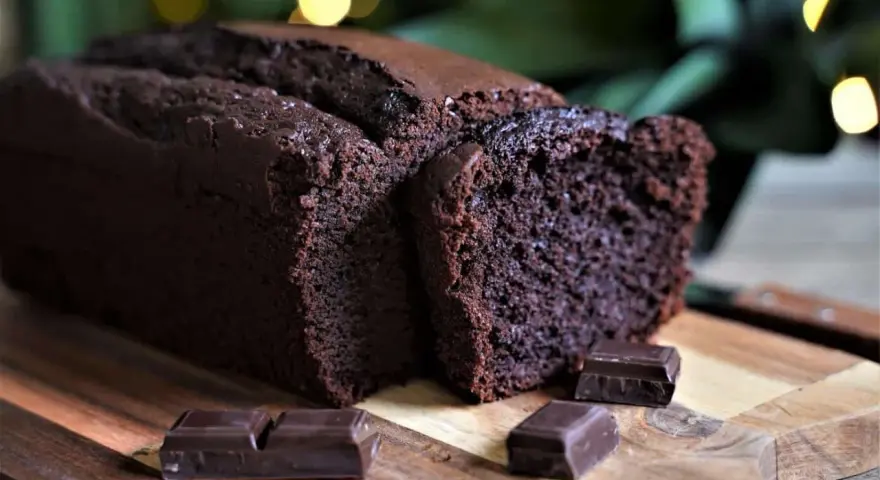 Un délicieux cake au chocolat, moelleux et intense, coupé en tranches sur une planche en bois, avec des carrés de chocolat noir en garniture.